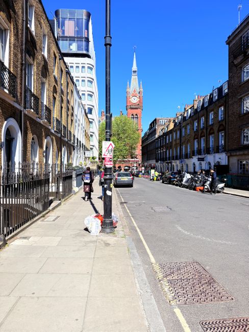 A wide city street leading towards a prominent red-brick clock tower with a pointed spire, situated in the background under a clear blue sky. On the left side of the image, there is a row of residenti